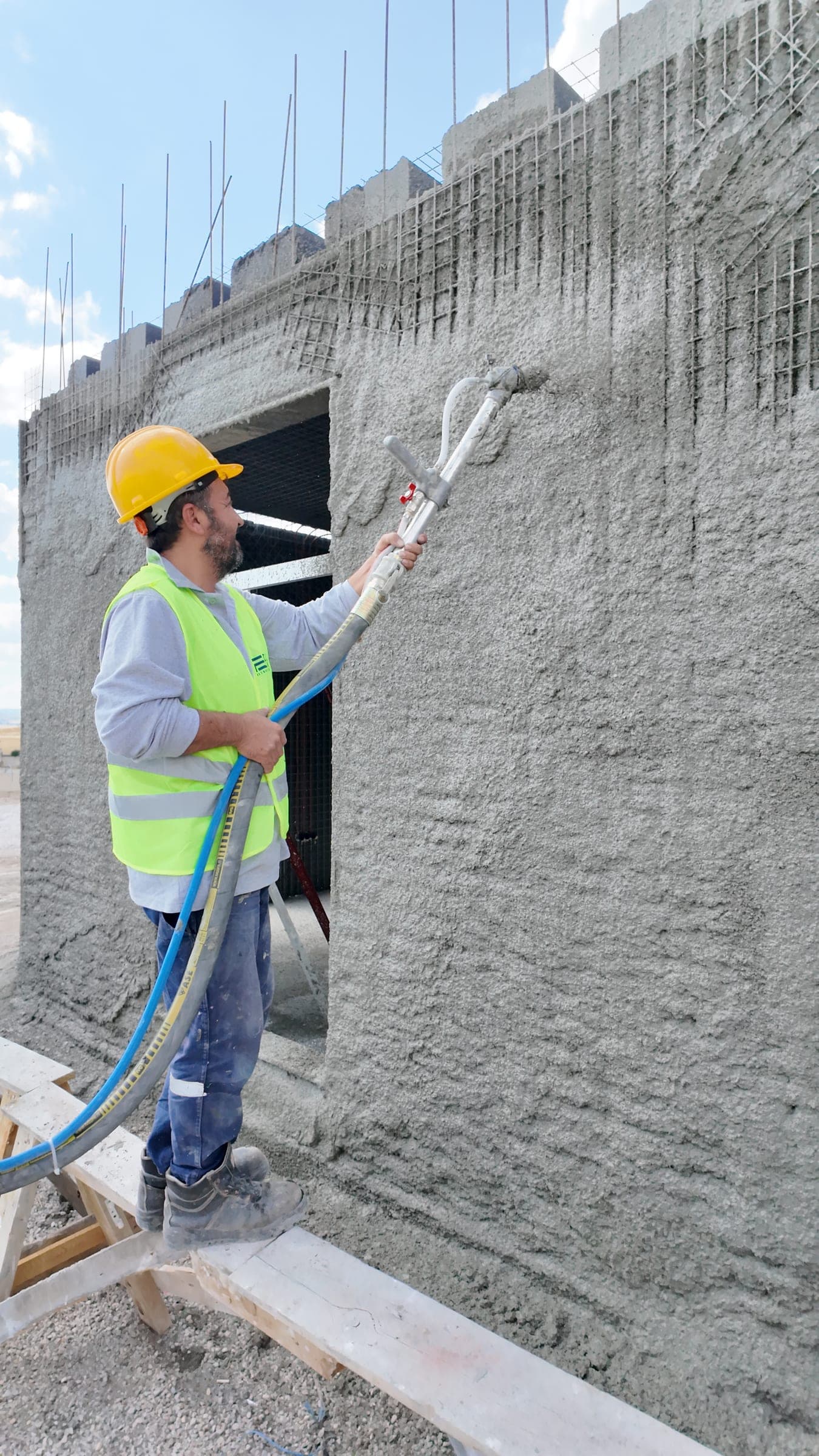 Bir işçi, çelik hasırlı panel duvarına püskürtme beton uyguluyor; taze yüzey pürüzlü ve gri.